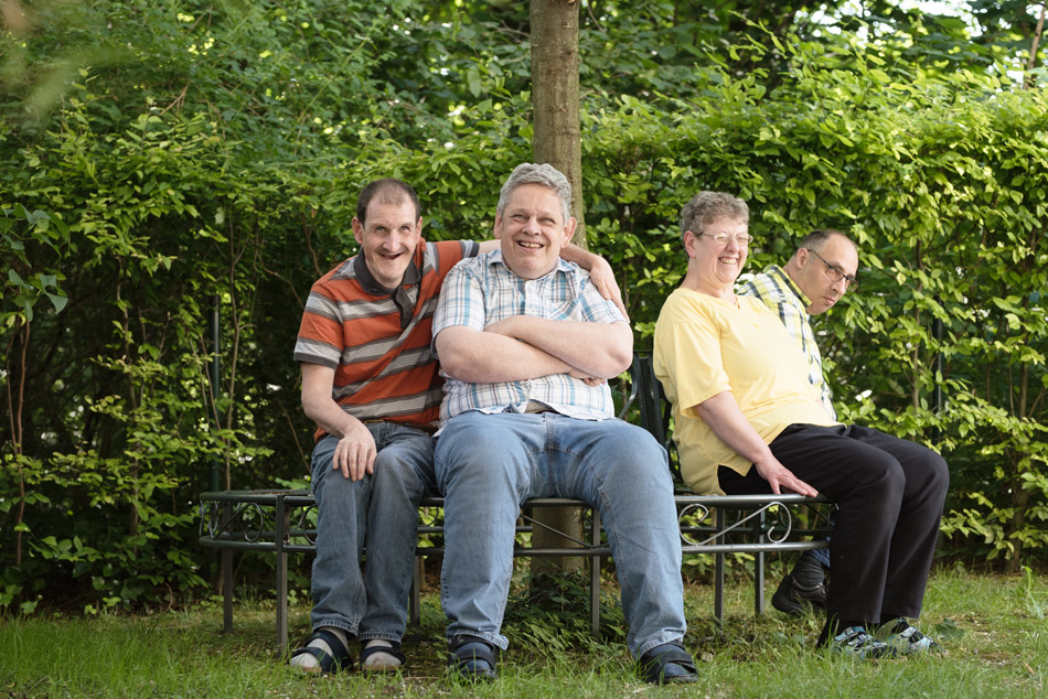 Vier Menschen mit Behinderung sitzen auf einer Bank um einen Baum herum und lachen.