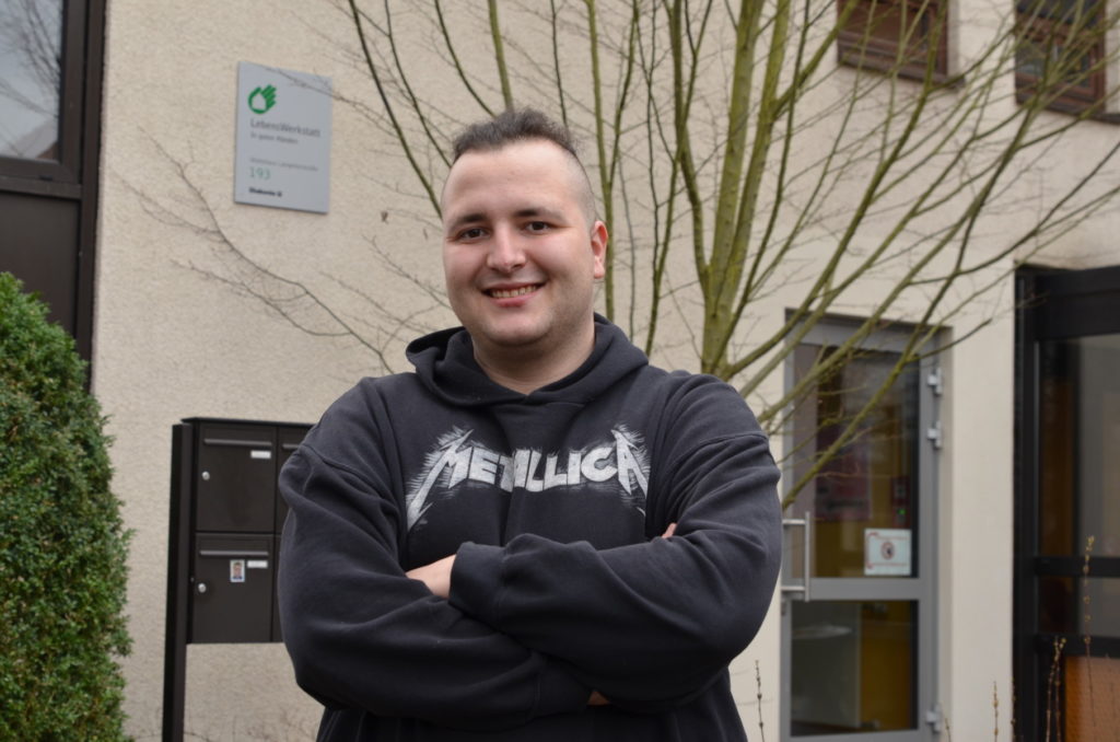 Junger Mann lächelt, trägt Metallica-Pullover, steht vor Gebäude mit Schild der LebensWerkstatt in modernem Stadtviertel.