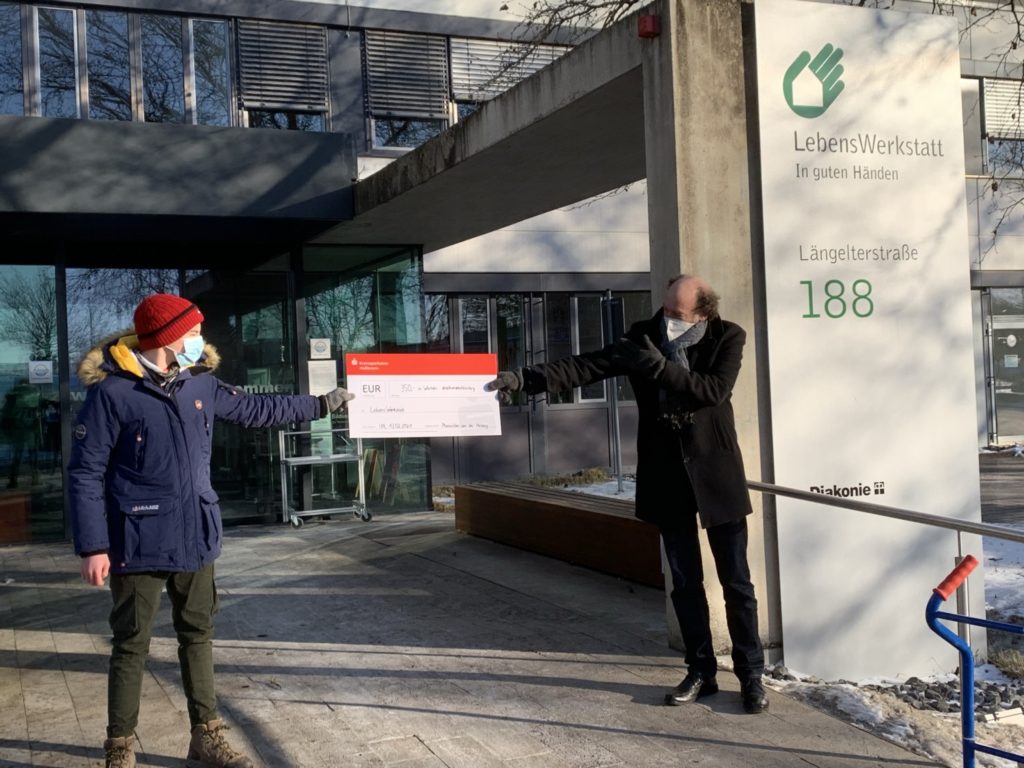 Zwei Personen übergeben symbolischen Scheck vor LebensWerkstatt Gebäude in der Längelterstraße 188.
