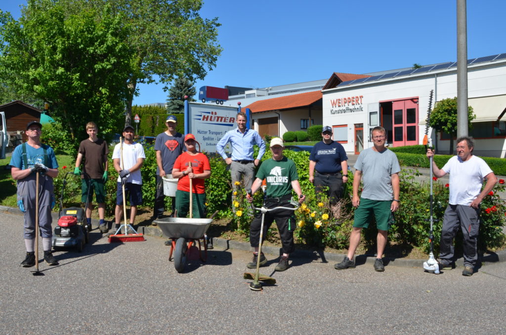Gruppe von acht Personen posiert mit Gartengeräten vor Firmengebäude und grüner Hecke, sonniger Tag.