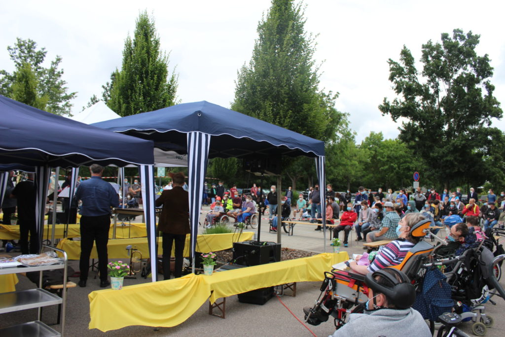 Versammlung im Freien mit Menschen unter Pavillons; viele sitzen und tragen Masken.