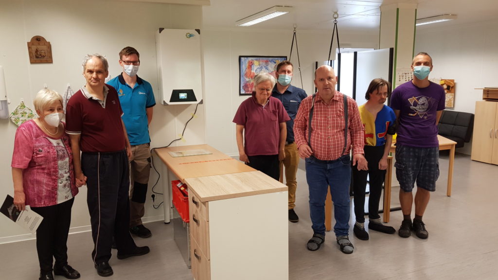 Eine Gruppe von Menschen mit Masken steht in einem Raum mit Holzmöbeln, lächelnd in die Kamera.