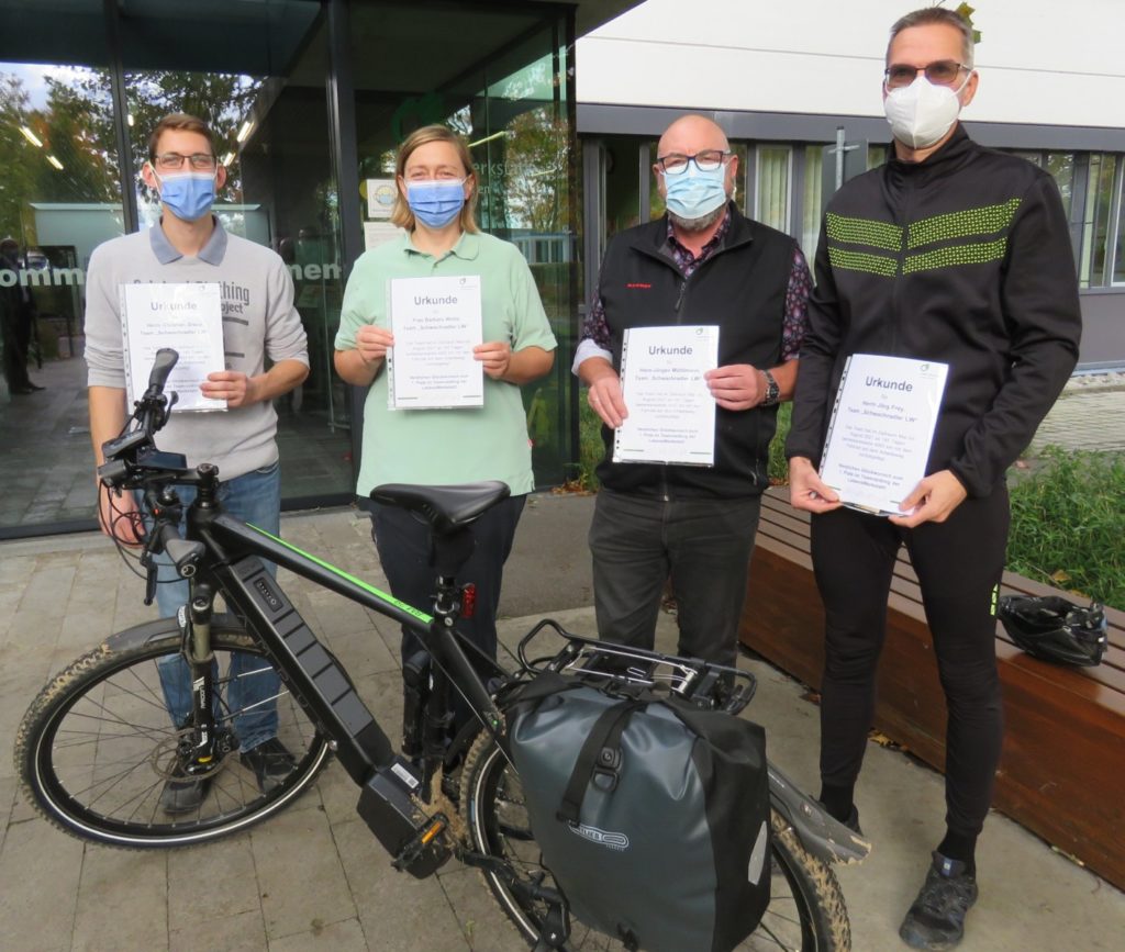 Vier Personen mit Masken halten Urkunden und stehen neben einem Fahrrad vor einem Gebäude.