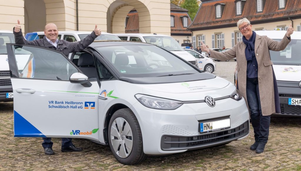 Zwei Personen freuen sich über ein VR Bank Elektroauto. Daumen hoch, vor einem historischen Gebäude.