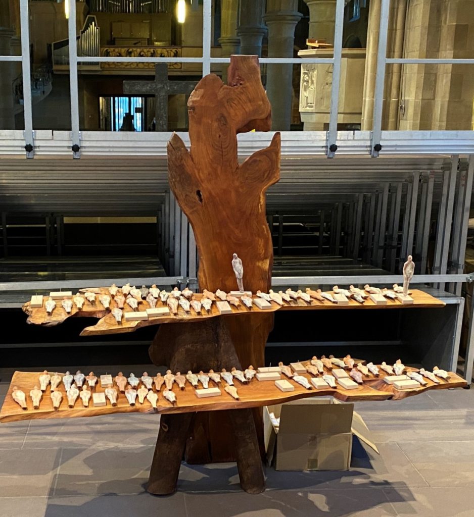 Holztisch in Kirche mit Holz- und Steinfiguren dekoriert, vor Metallstruktur und Kirchenfenster im Hintergrund.