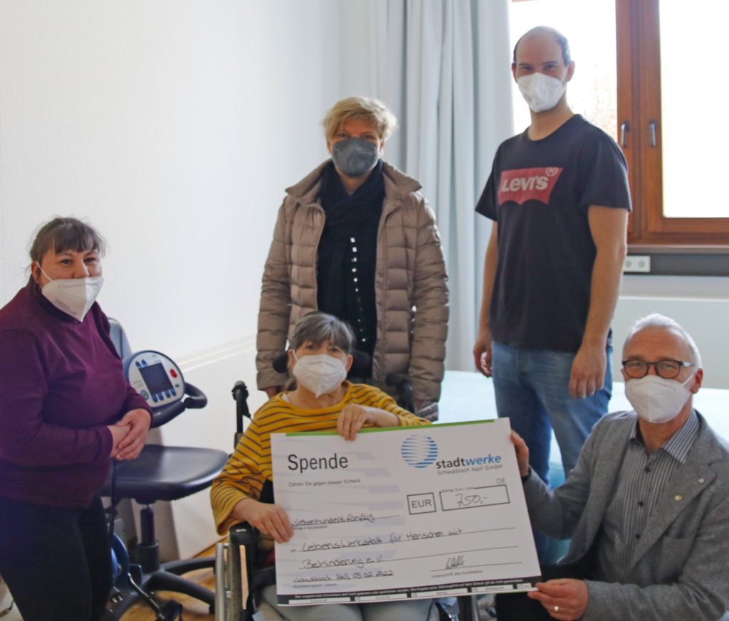 Gruppe mit großem Spenden-Scheck in einem Raum, alle tragen Masken und zeigen Unterstützung.