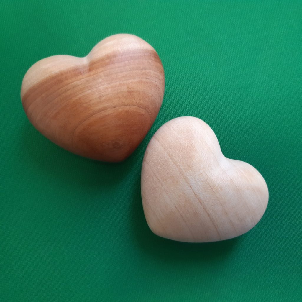 Zwei holzfarbene Herzfiguren auf grünem Hintergrund aus Holz gefertigt, liegen nebeneinander.