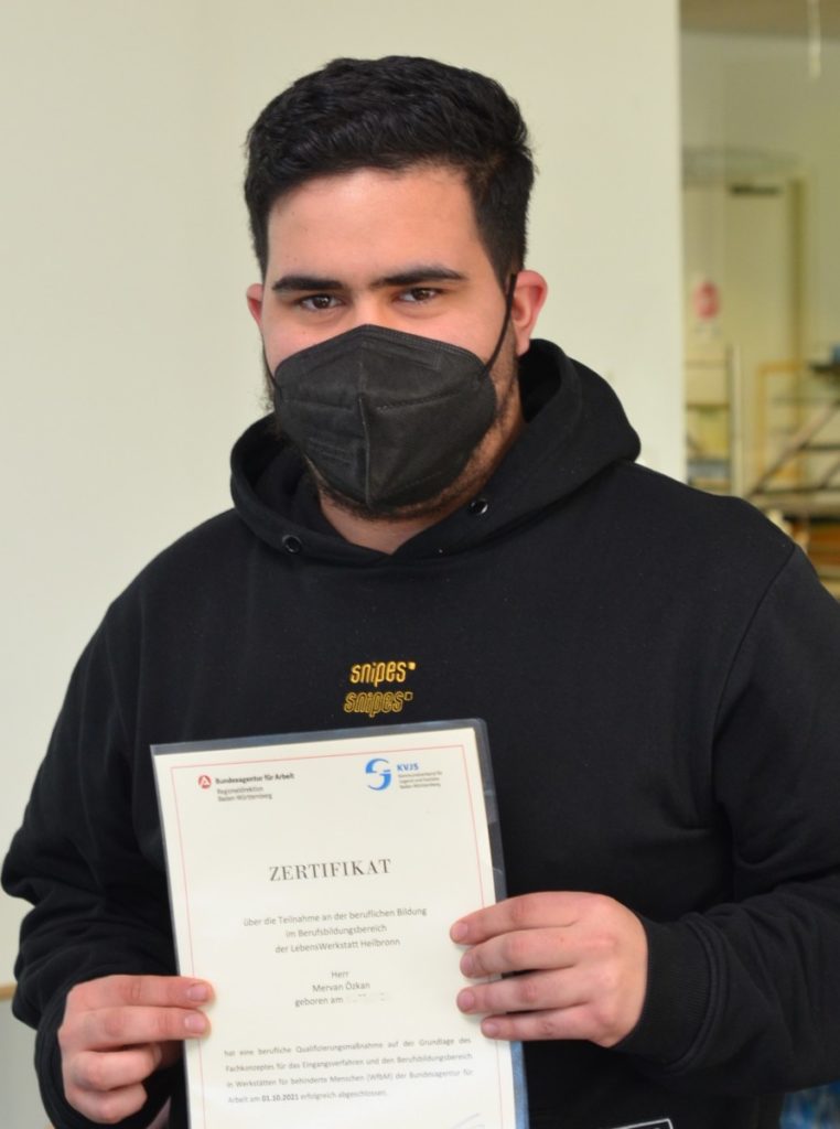 Mann mit schwarzer Maske hält ein Zertifikat der Bundesagentur für Arbeit in der Hand, schaut direkt in die Kamera.