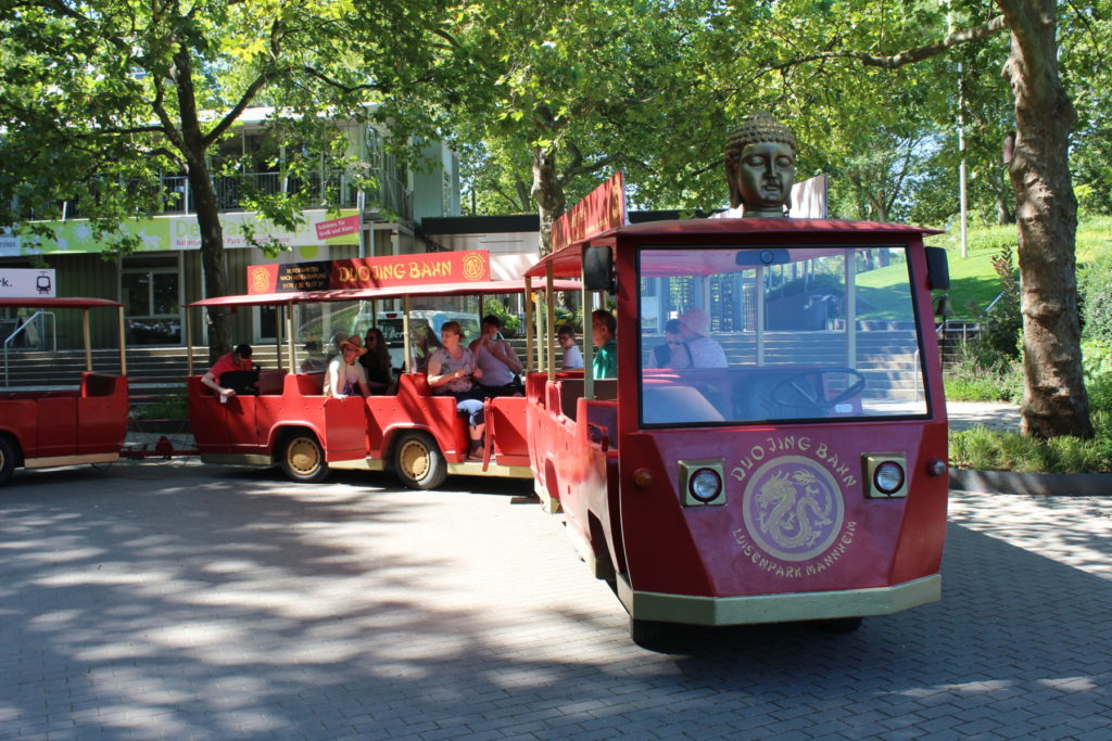 Roter Parkzug Duo Jing Bahn mit Buddha-Kopf, fährt durch sonnigen Park, Fahrgäste an Bord.