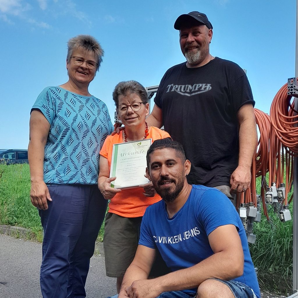 Vier Menschen posieren draußen mit einer Urkunde, lächeln in die Kamera und zeigen Stolz.