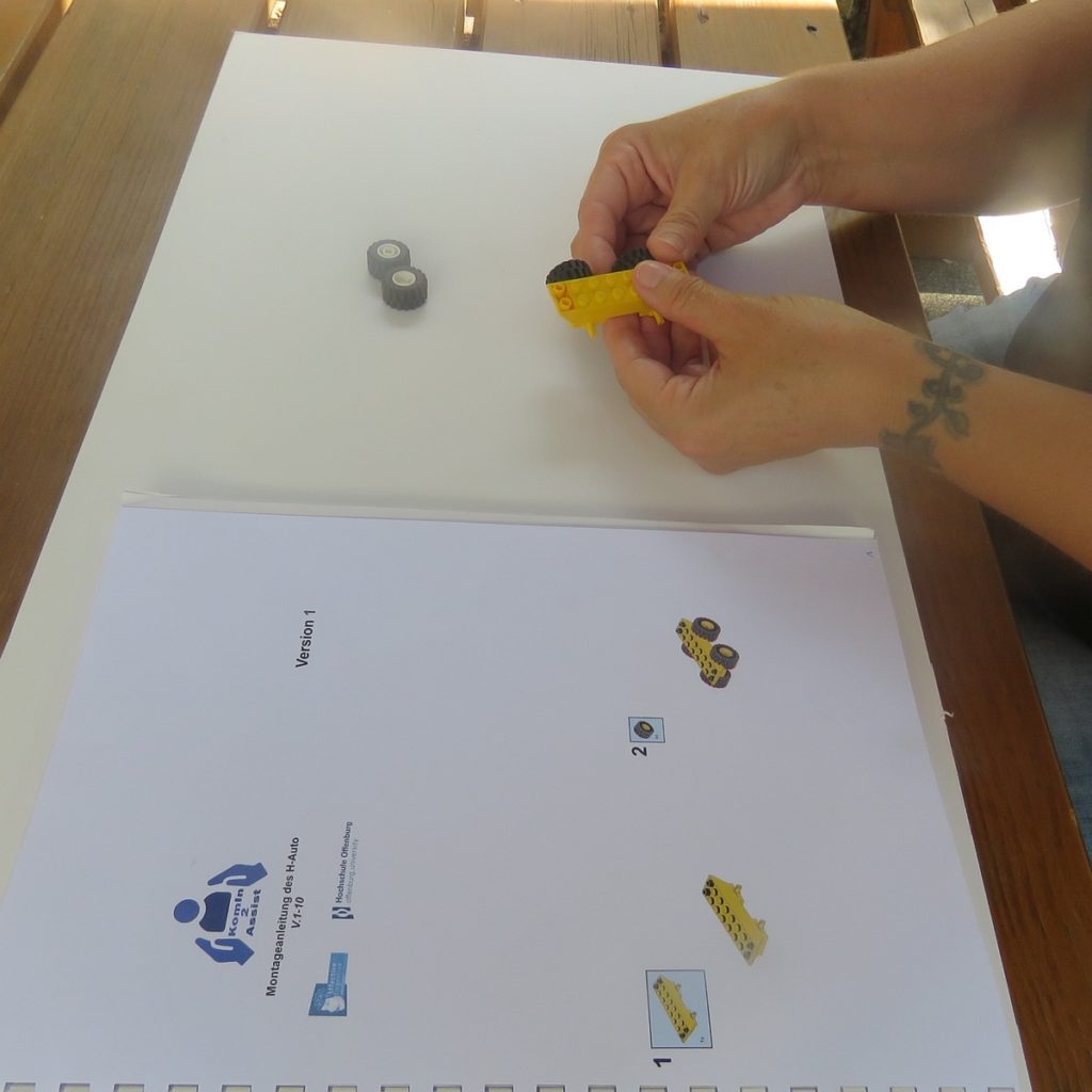 Person baut ein kleines Modellauto aus Lego mit Anleitung auf weißem Tisch zusammen.