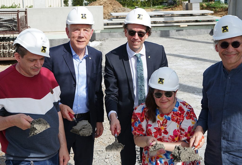 Fünf Personen mit Bauhelmen und Maurerkellen bei einer Grundsteinlegung auf einer Baustelle.