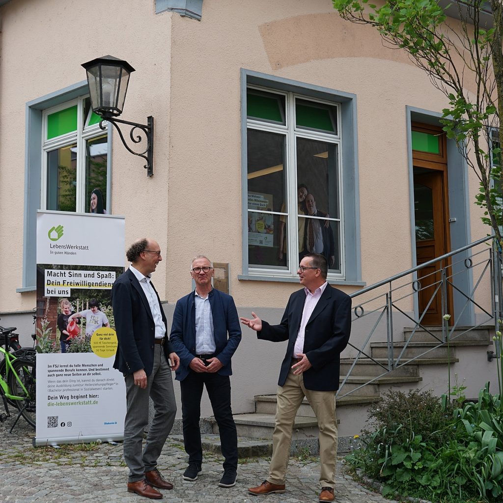 Drei Männer stehen vor einem Gebäude der LebensWerkstatt und unterhalten sich neben einem Banner über Freiwilligendienste.