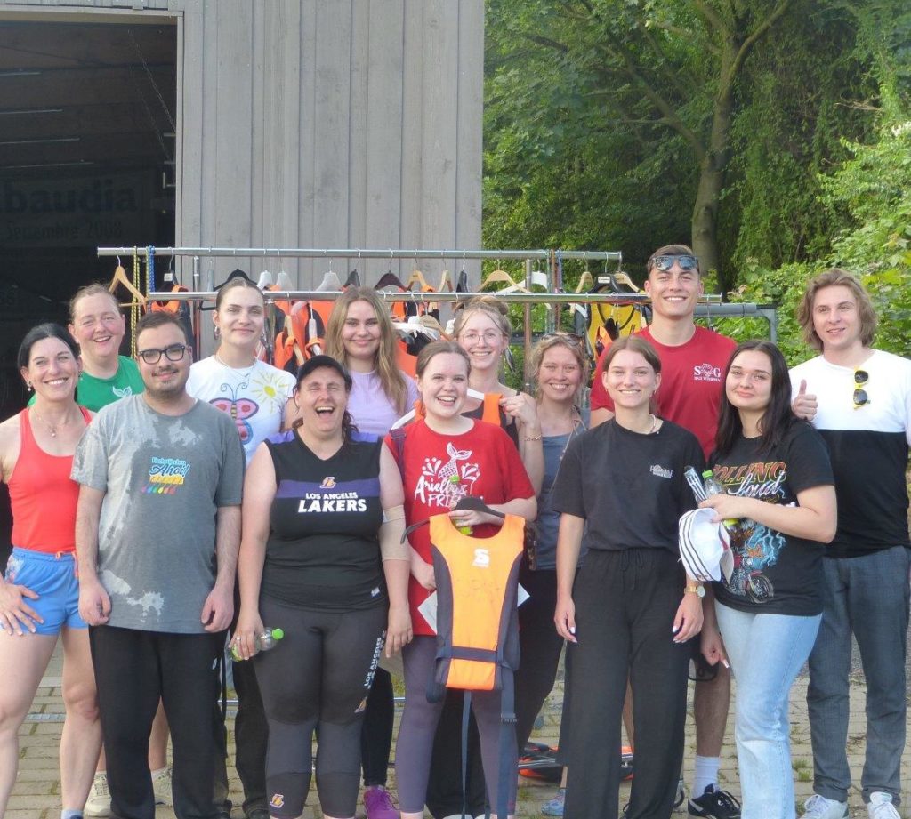 Eine Gruppe von Menschen lächelt draußen vor einer Hütte mit Rettungswesten im Hintergrund.