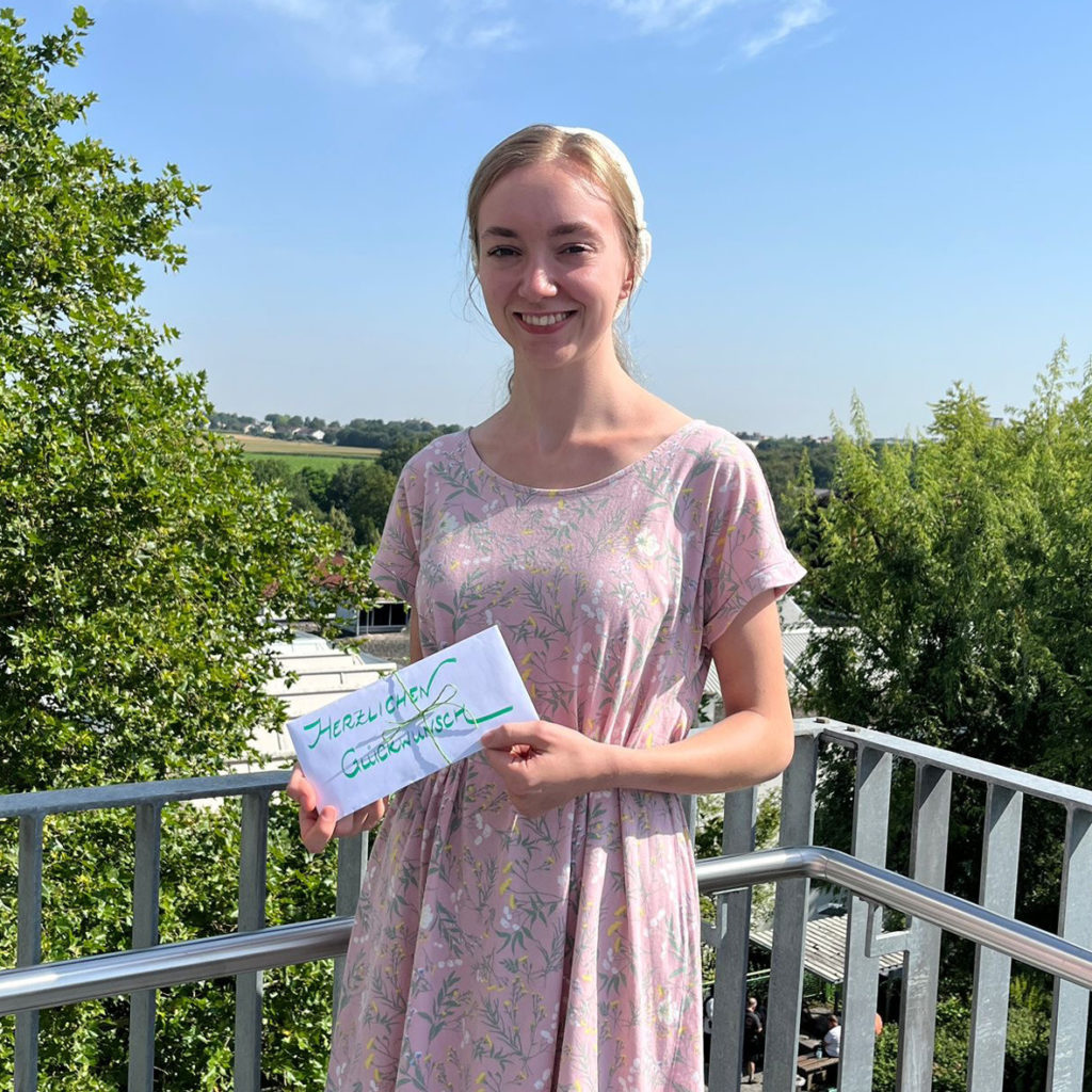 Junge Frau im Sommerkleid hält eine Grußkarte mit der Aufschrift Herzlichen Glückwunsch auf einem Balkon.