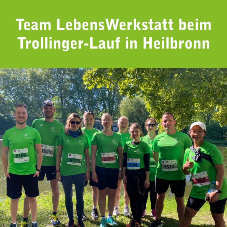 Gruppe von zehn Läuferinnen und Läufern des Teams „LebensWerkstatt“ beim Trollinger-Lauf in Heilbronn. Alle tragen grüne Laufshirts mit dem Logo der LebensWerkstatt und stehen bei sonnigem Wetter im Grünen vor einem Fluss. Oben im Bild steht der Text: „Team LebensWerkstatt beim Trollinger-Lauf in Heilbronn“.