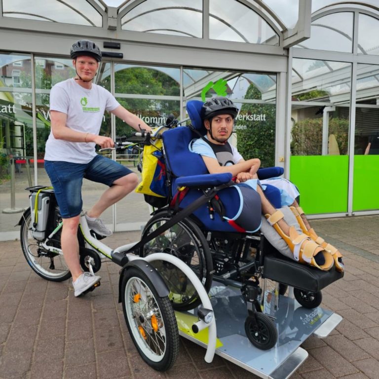 ein mann sitzt auf einem fahrrad das vornei einen aufbau hat in welchem ein anderer mann in einem rollstuhl sitzt
