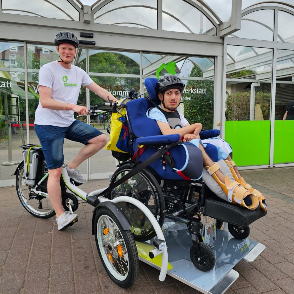 ein mann sitzt auf einem fahrrad das vornei einen aufbau hat in welchem ein anderer mann in einem rollstuhl sitzt