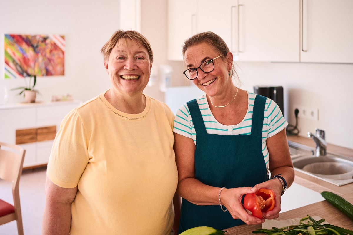 Zwei lächelnde Frauen in einer Küche, die zusammen Gemüse zubereiten.