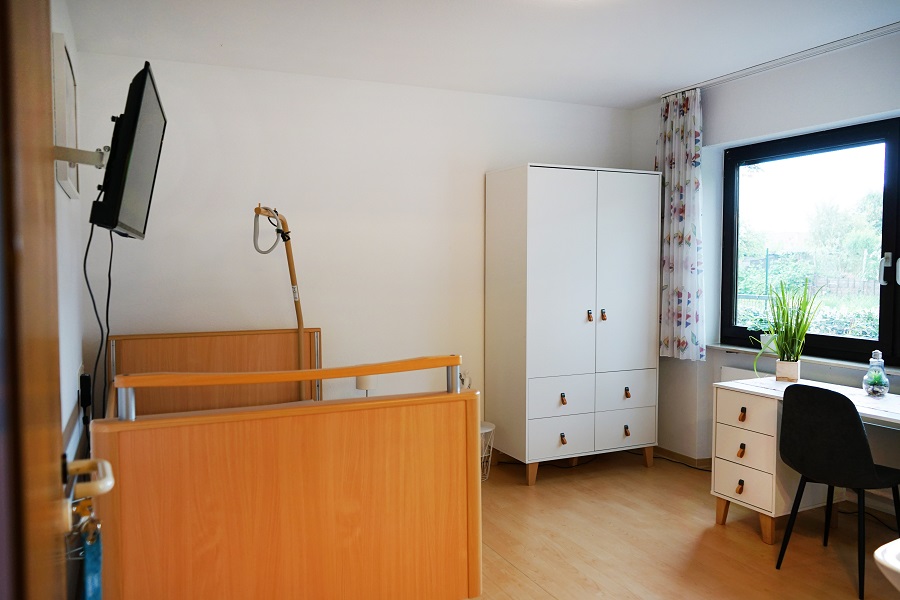 Helles Schlafzimmer mit Pflegebett, Schrank, Schreibtisch und Fenster mit Ausblick ins Grüne, freundliche Einrichtung.