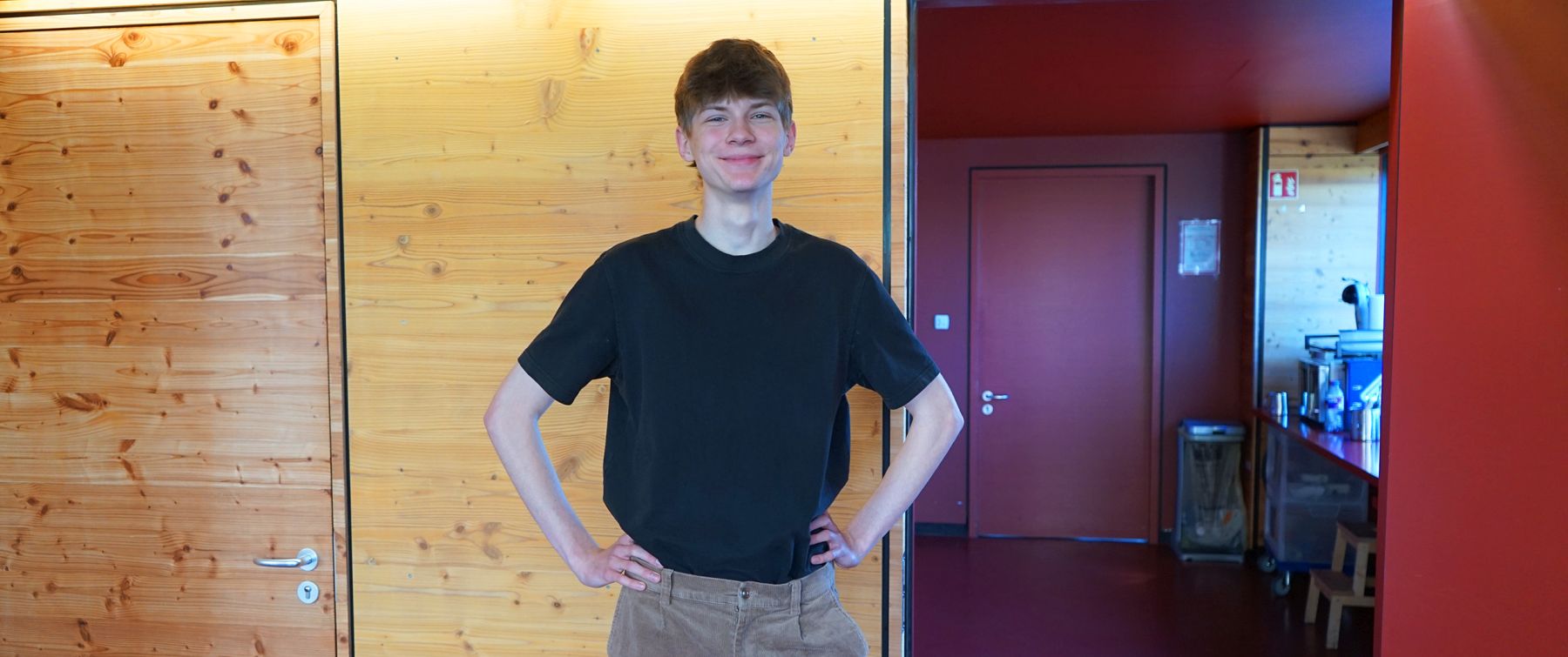 Person in schwarzem T-Shirt lächelt und steht vor einer Holzwand in einem Raum mit roten Türen.