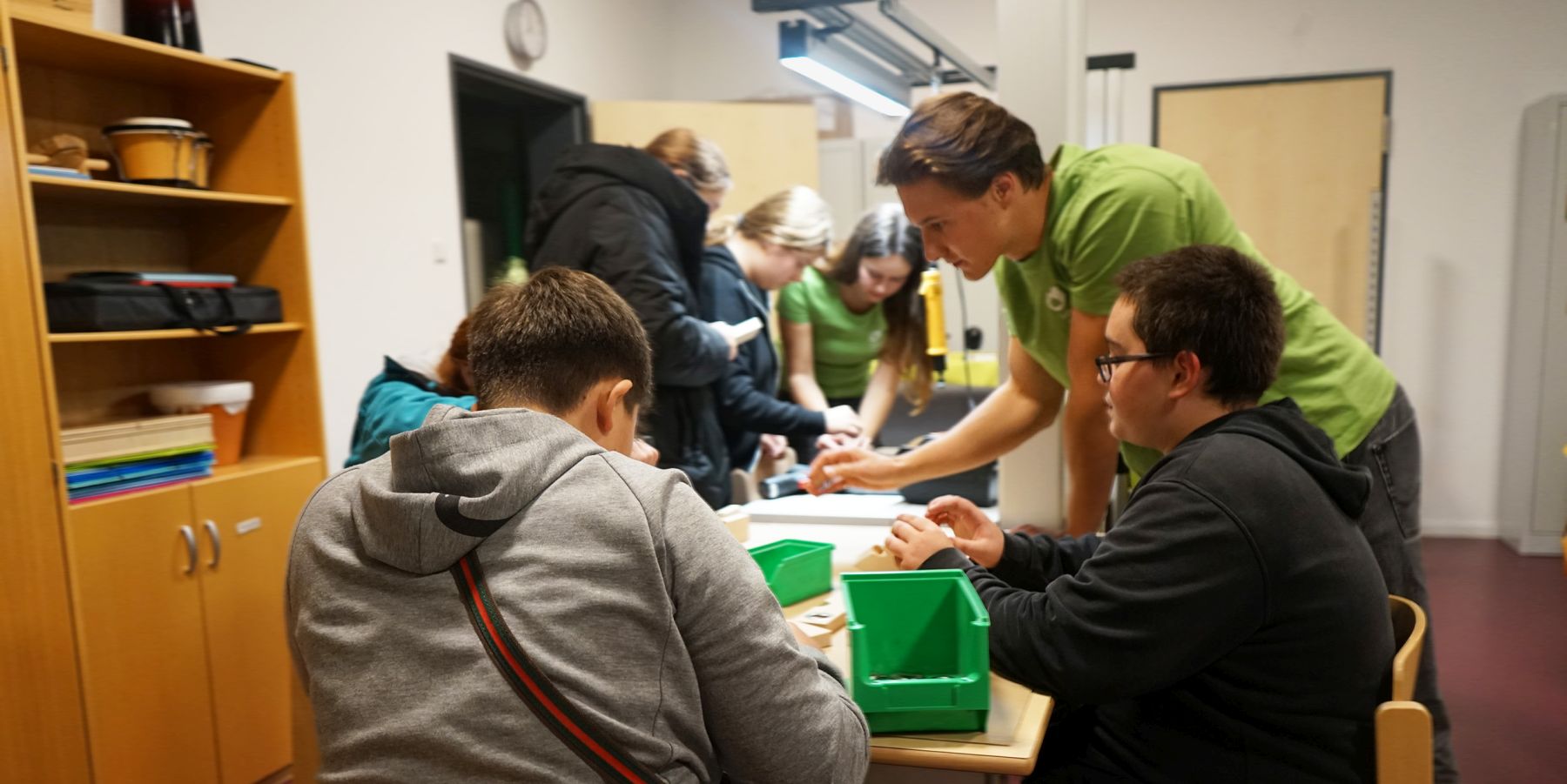 Menschen arbeiten zusammen in einem Klassenzimmer an einem Tisch mit grünen Boxen, helfen einander bei Aufgaben.