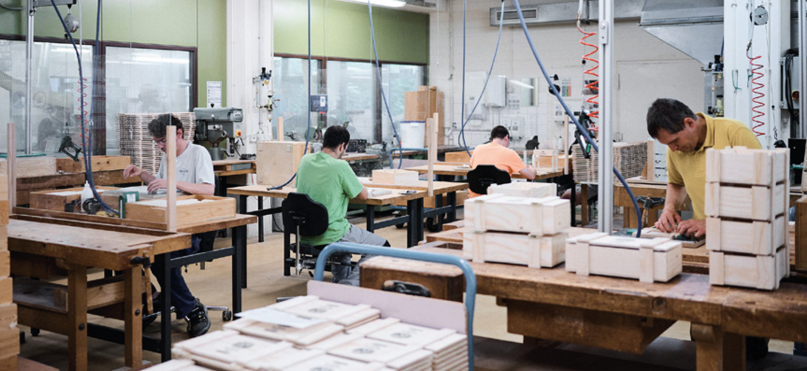 Arbeiter in einer Holzwerkstatt mit Tischen, Werkzeugen und Holzstapeln. Produktions- oder Handwerksumgebung.