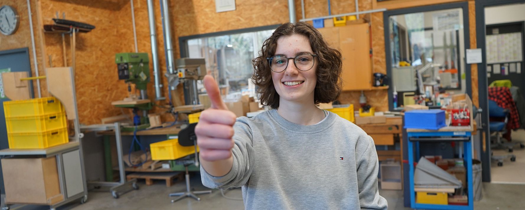 Ein junger Mensch steht in einer modernen Werkstatt und hebt den Daumen hoch