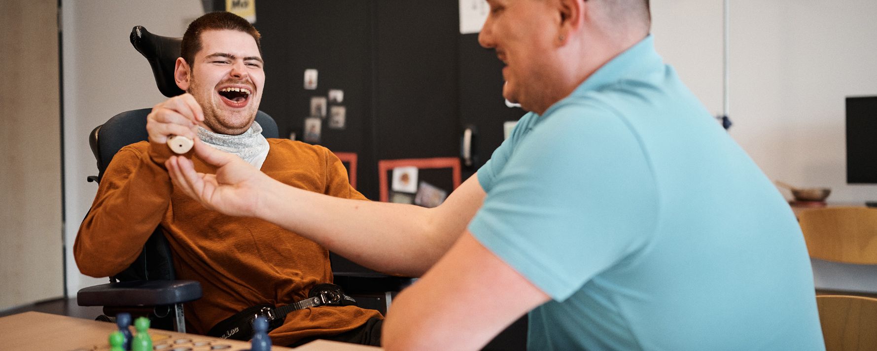 Zwei Männer spielen freudig ein Brettspiel, einer im Rollstuhl, lachend in entspannter Atmosphäre.