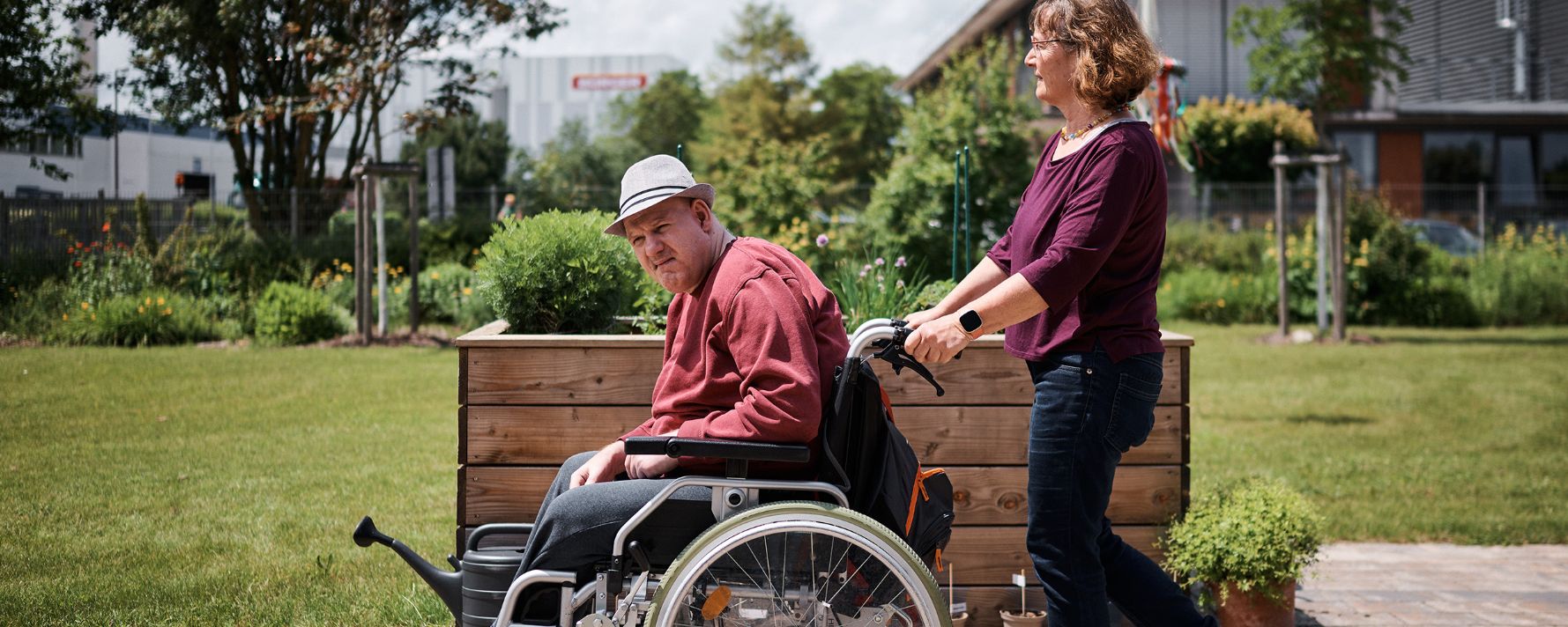 Frau schiebt Mann im Rollstuhl durch einen sonnigen Garten; beide lächeln.