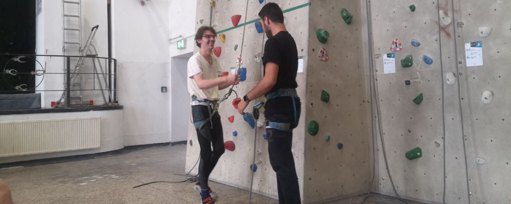 Zwei Personen in Kletterkleidung an einer Indoor-Kletterwand, die sich auf das Klettern vorbereiten.