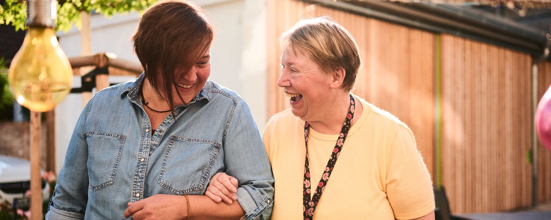 Zwei lachende Menschen gehen Arm in Arm im Freien, umgeben von Lichtern und Bäumen.