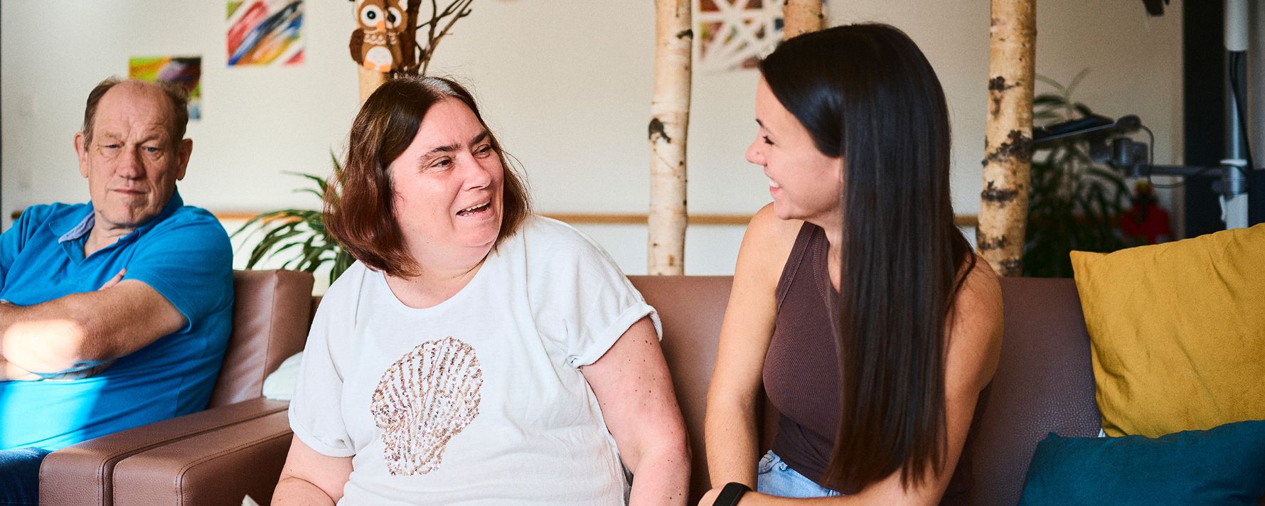 Drei Personen auf einem Sofa, lächelnd und im Gespräch, gemütliche Umgebung mit Pflanzen.