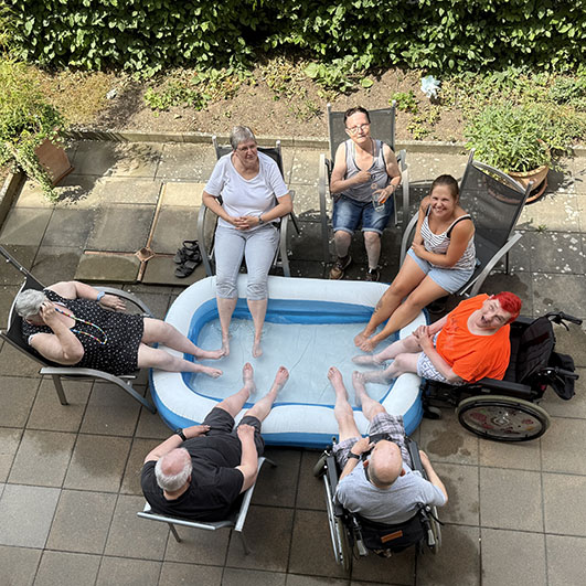 Sechs Personen sitzen entspannt um ein aufblasbares Planschbecken im Garten und genießen den Tag.