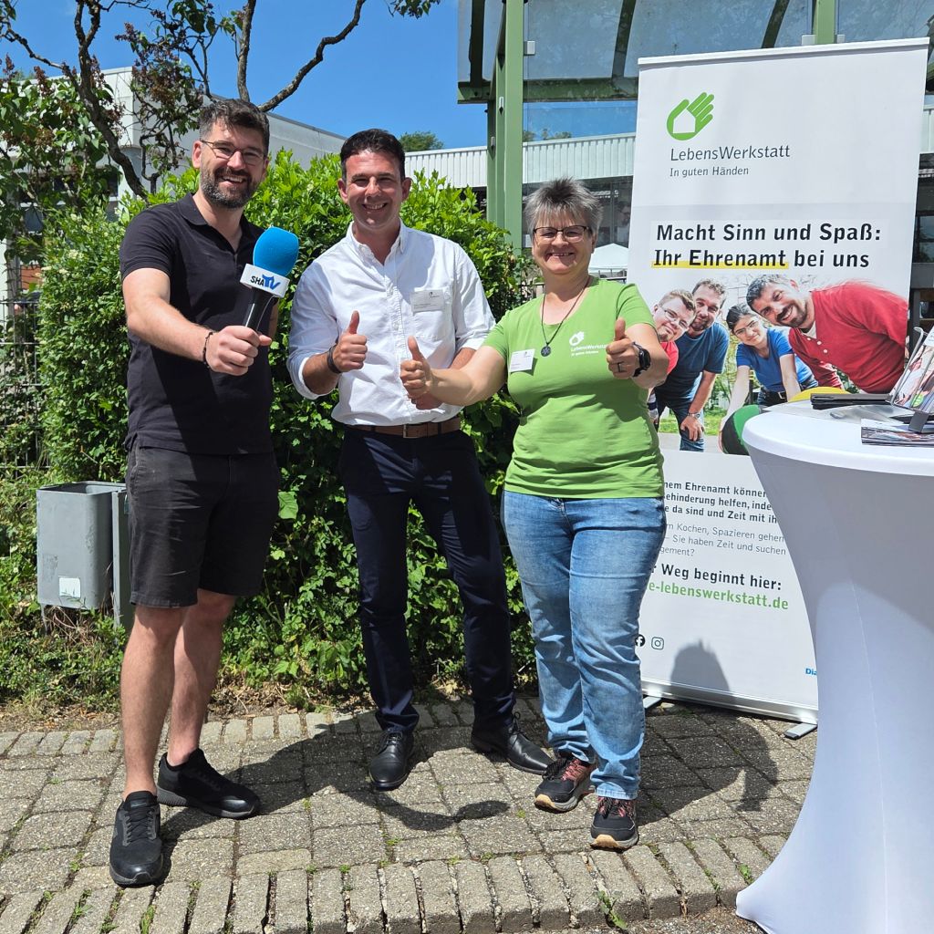 Drei Personen geben Daumen hoch und stehen vor einem Informationsstand über Ehrenamt. Es ist ein sonniger Tag.