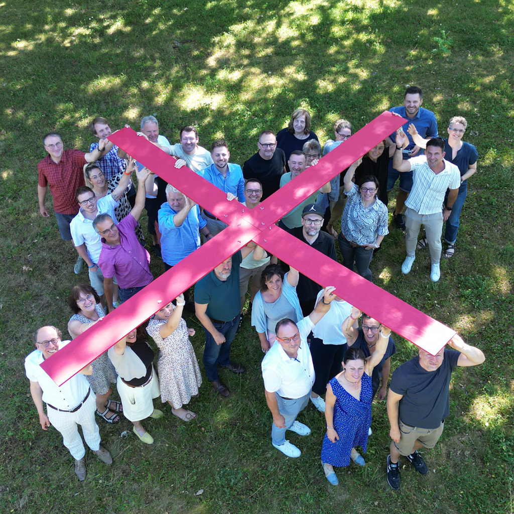 Gruppe von Menschen hält ein großes rotes X über ihren Köpfen auf einer Wiese an einem sonnigen Tag.