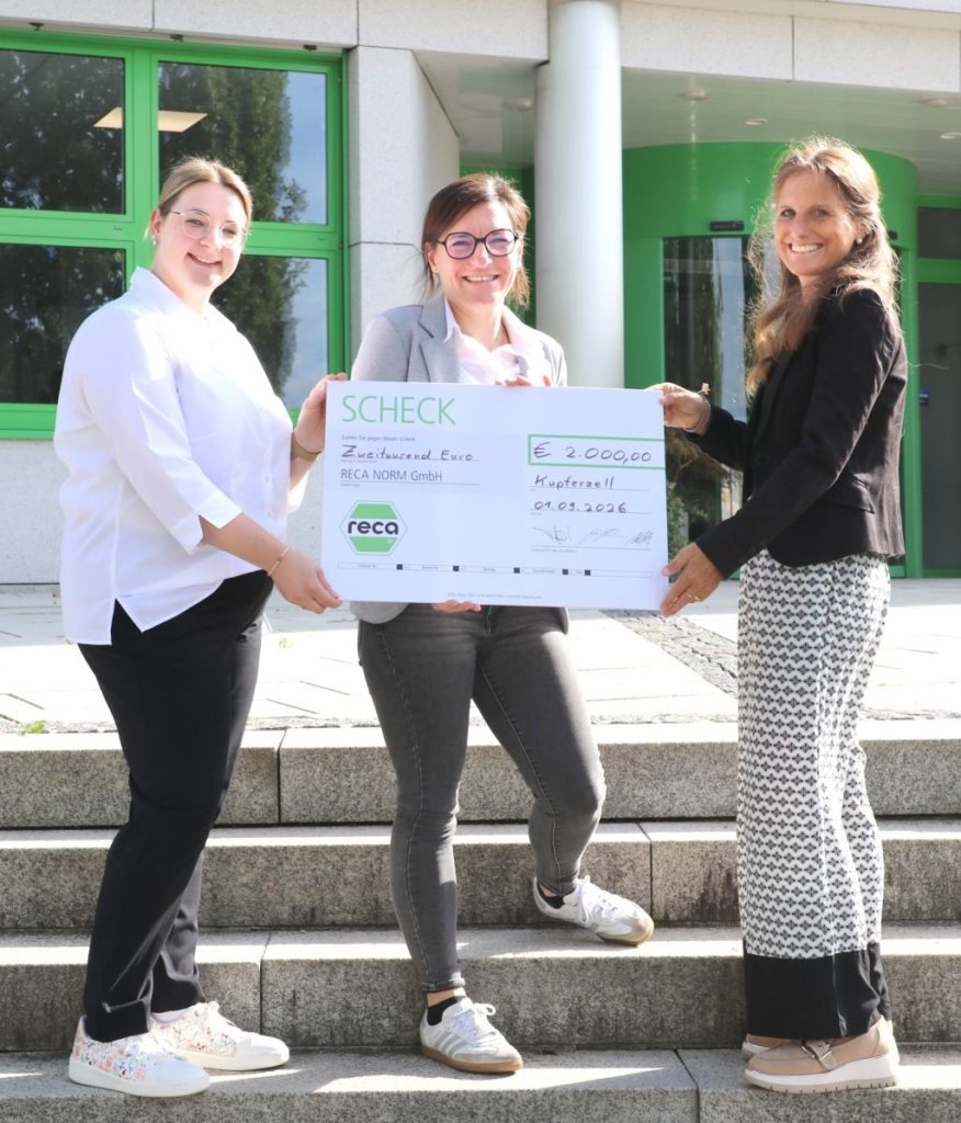 Drei Frauen halten einen großen Scheck über 2000 Euro vor einem Gebäude von RECA NORM GmbH.