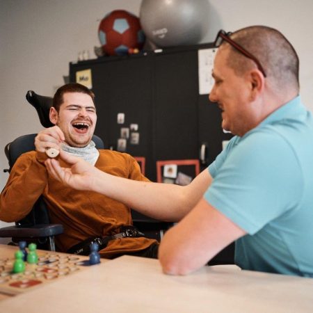 Ein junger Mann lacht glücklich, während er mit einem anderen Mann ein Spiel spielt. Sie sitzen an einem Tisch.