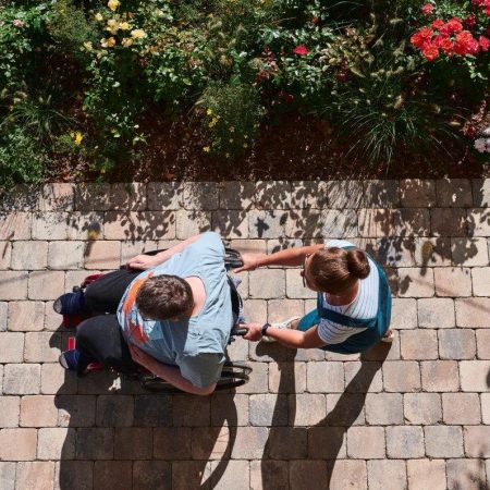 Eine Person im Rollstuhl wird von einer anderen Person auf einem gepflasterten Weg mit Blumen begleitet.