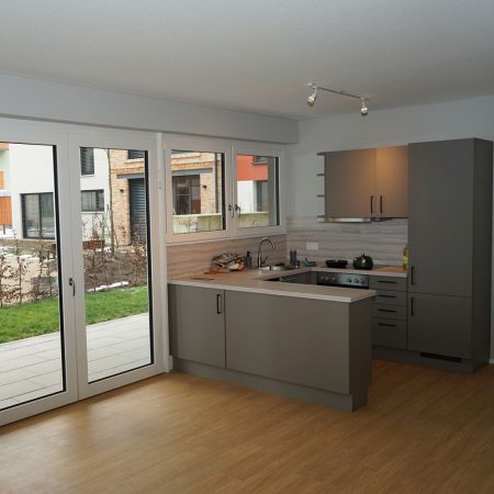 Moderne Küche mit grauen Schränken, großen Fenstern und Blick ins Freie. Helles, einladendes Ambiente.