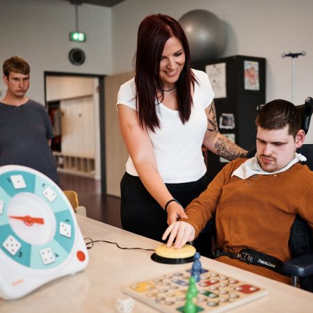Fachkraft unterstützt Mann im Rollstuhl bei Aktivität mit großem Knopf am Tisch, im Hintergrund steht eine weitere Person.