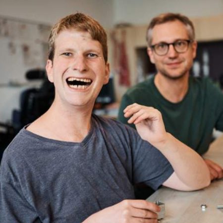 Mensch in grauem T-Shirt lächelt, sitzt an Tisch, eine weitere Person unscharf im Hintergrund.